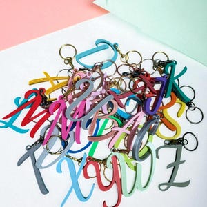 May include: A collection of colorful letter keychains in various hues, including red, blue, green, and pink. Each keychain features a different letter of the alphabet and includes a gold-colored keyring. The keychains are arranged on a white surface.