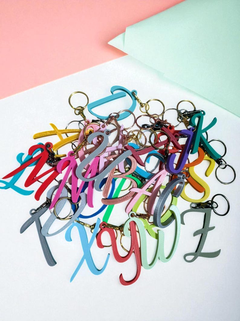 May include: A collection of colorful letter keychains in various hues, including red, blue, green, and pink. Each keychain features a different letter of the alphabet and includes a gold-colored keyring. The keychains are arranged on a white surface.