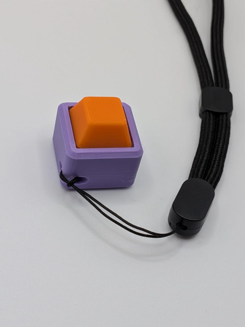 May include: A purple square-shaped object with an orange square button on top. A black lanyard is attached to the object. The lanyard has a black plastic clasp and a black cord.