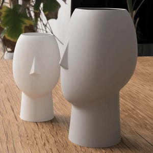 May include: Two white ceramic vases shaped like human heads. The vases have a minimalist design with a smooth, matte finish. One vase is smaller than the other. The vases are placed on a wooden surface.