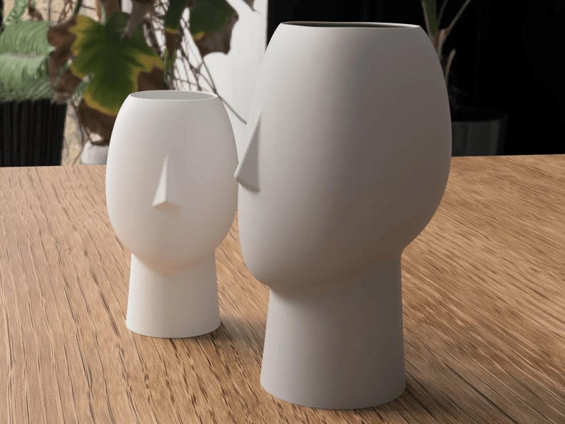 May include: Two white ceramic vases shaped like human heads. The vases have a minimalist design with a smooth, matte finish. One vase is smaller than the other. The vases are placed on a wooden surface.