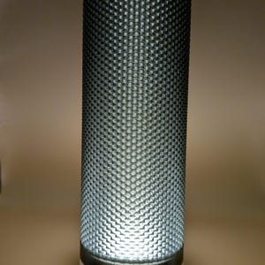 K&ouml;nnte beinhalten: Zylindrische Lampe mit strukturierter, dunkelgrauer Au&szlig;enseite. Die Lampe hat einen wei&szlig;en Sockel und ein helles Licht, das von innen ausgeht. Ein wei&szlig;es Netzkabel erstreckt sich von der Basis. Das Design der Lampe weist ein sich wiederholendes Muster aus kleinen, erhabenen Quadraten auf.