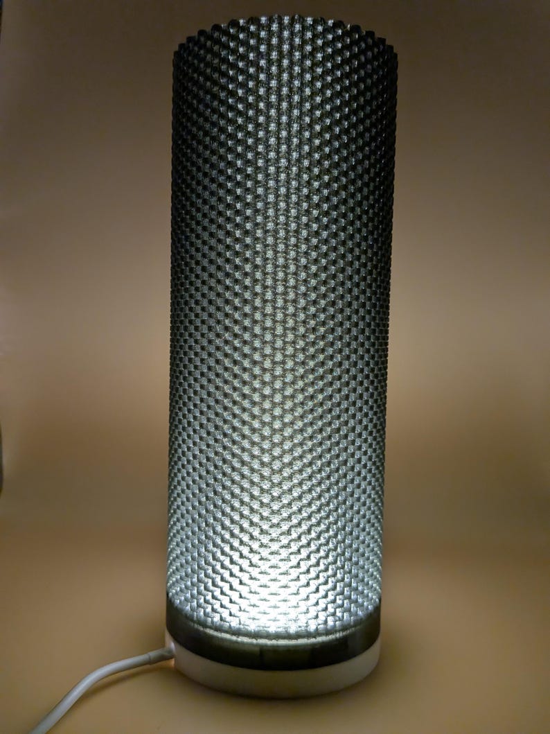 K&ouml;nnte beinhalten: Zylindrische Lampe mit strukturierter, dunkelgrauer Au&szlig;enseite. Die Lampe hat einen wei&szlig;en Sockel und ein helles Licht, das von innen ausgeht. Ein wei&szlig;es Netzkabel erstreckt sich von der Basis. Das Design der Lampe weist ein sich wiederholendes Muster aus kleinen, erhabenen Quadraten auf.