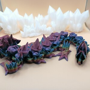 May include: A 3D-printed sculpture of a dragon-like creature with rose and crystal elements. The dragon features a gradient of colors, including purple, blue, and green. White crystal formations are in the background.