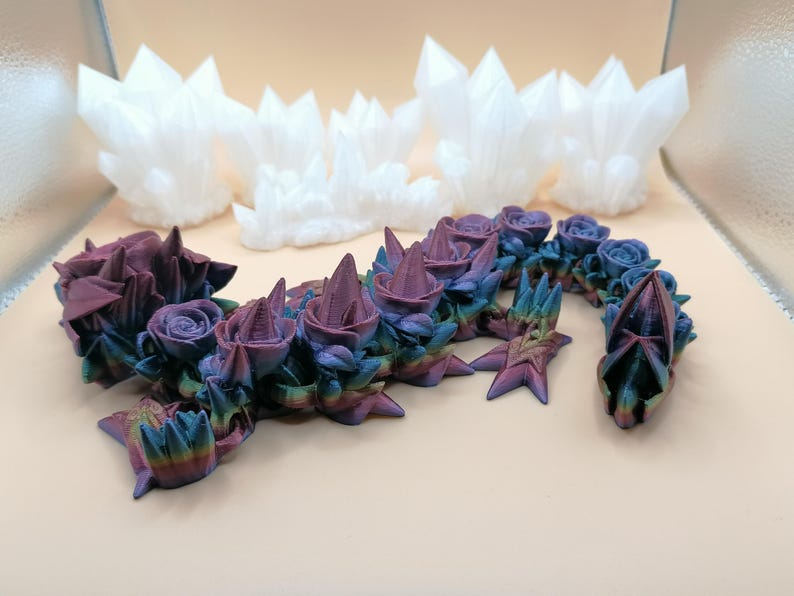 May include: A 3D-printed sculpture of a dragon-like creature with rose and crystal elements. The dragon features a gradient of colors, including purple, blue, and green. White crystal formations are in the background.