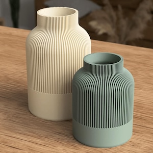 Puede incluir: Dos jarrones decorativos con un dise&ntilde;o acanalado. Uno es de color blanquecino y el otro es de color verde azulado apagado. Ambos jarrones tienen una base de color s&oacute;lido y se exhiben sobre una superficie de madera. Los jarrones son cil&iacute;ndricos con un cuello estrecho.