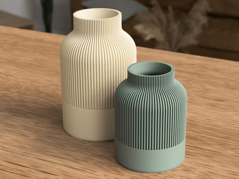 Puede incluir: Dos jarrones decorativos con un dise&ntilde;o acanalado. Uno es de color blanquecino y el otro es de color verde azulado apagado. Ambos jarrones tienen una base de color s&oacute;lido y se exhiben sobre una superficie de madera. Los jarrones son cil&iacute;ndricos con un cuello estrecho.