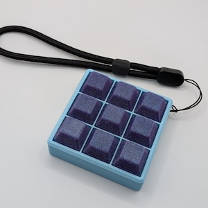 May include: A light blue square fidget toy with nine purple square buttons. The buttons have a glittery finish. A black lanyard is attached to the toy.