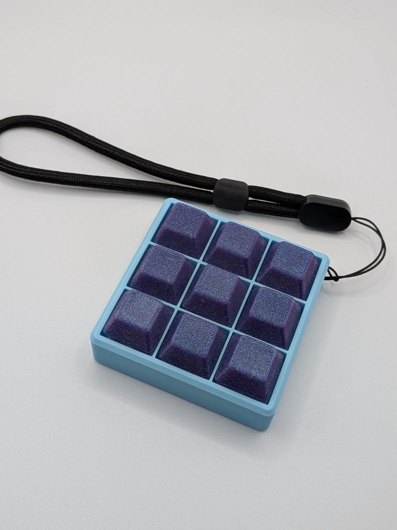 May include: A light blue square fidget toy with nine purple square buttons. The buttons have a glittery finish. A black lanyard is attached to the toy.