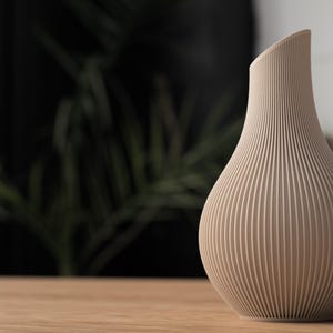May include: A beige, ribbed vase with a curved, asymmetrical top. The vase has a bulbous base and tapers upwards, with vertical lines creating a textured surface. The vase is sitting on a wooden surface.