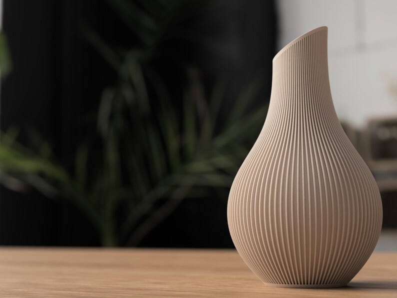 May include: A beige, ribbed vase with a curved, asymmetrical top. The vase has a bulbous base and tapers upwards, with vertical lines creating a textured surface. The vase is sitting on a wooden surface.