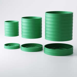 Op de afbeelding: Een set groene cilindervormige plantenbakken en bijpassende schotels. De plantenbakken vari&euml;ren in grootte, met horizontale geribbelde details. De schotels zijn plat en cirkelvormig. De objecten zijn felgroen van kleur en staan tegen een witte achtergrond.