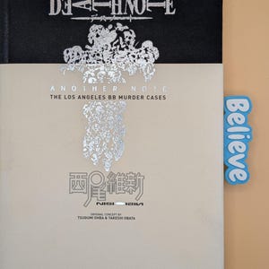 May include: A black hardcover book titled Death Note Another Note The Los Angeles BB Murder Cases with a beige dust jacket featuring silver foil text and graphics. A light blue rectangular tab with the word Believe is attached to the side.