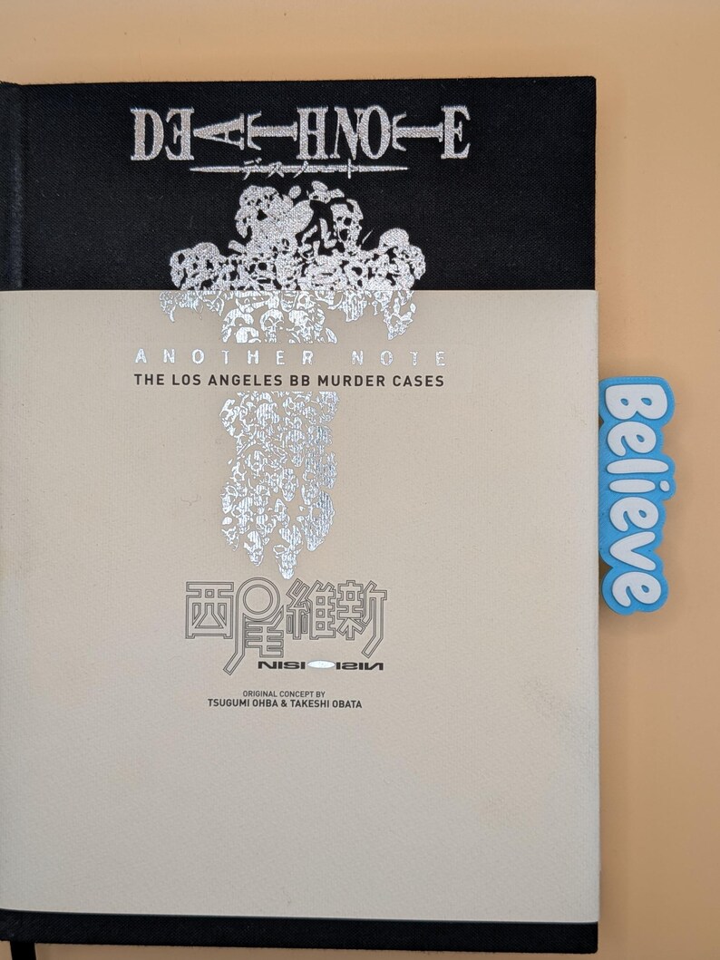 May include: A black hardcover book titled Death Note Another Note The Los Angeles BB Murder Cases with a beige dust jacket featuring silver foil text and graphics. A light blue rectangular tab with the word Believe is attached to the side.