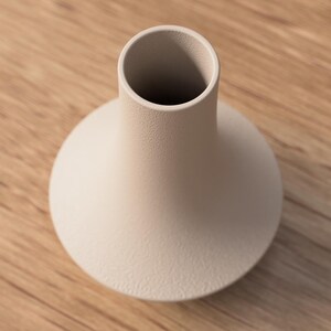 May include: A beige ceramic vase with a rounded base and a cylindrical neck. The vase has a textured surface and sits on a wooden surface. The vase is empty and has a simple, modern design.