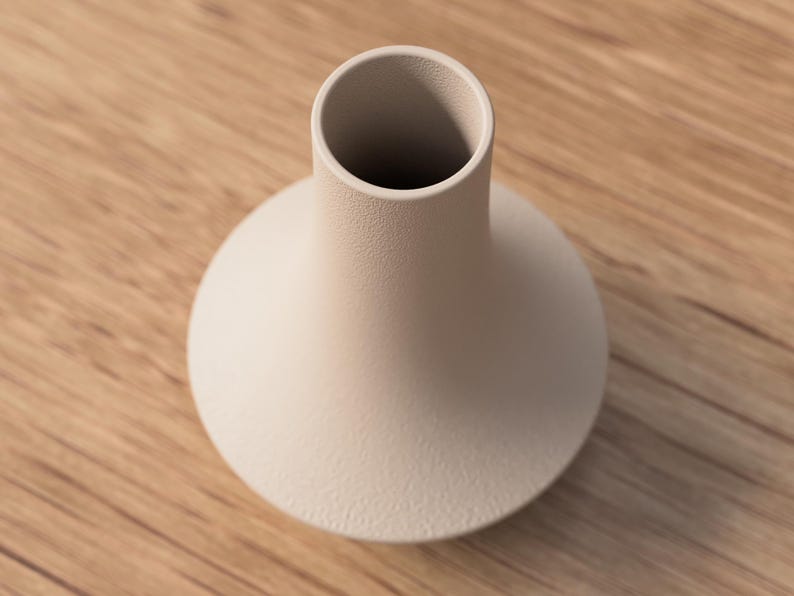 May include: A beige ceramic vase with a rounded base and a cylindrical neck. The vase has a textured surface and sits on a wooden surface. The vase is empty and has a simple, modern design.