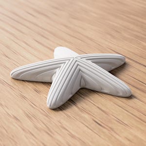 May include: A white, five-pointed starfish-shaped decorative object with ridged detailing. The object is resting on a light brown wooden surface, creating a contrast in textures and colors. The starfish design is a common theme in coastal decor.
