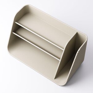 May include: A beige desk organizer with two shelves and a divided compartment. The organizer has a rounded shape with a textured side and a smooth, matte finish. It is designed to hold office supplies.
