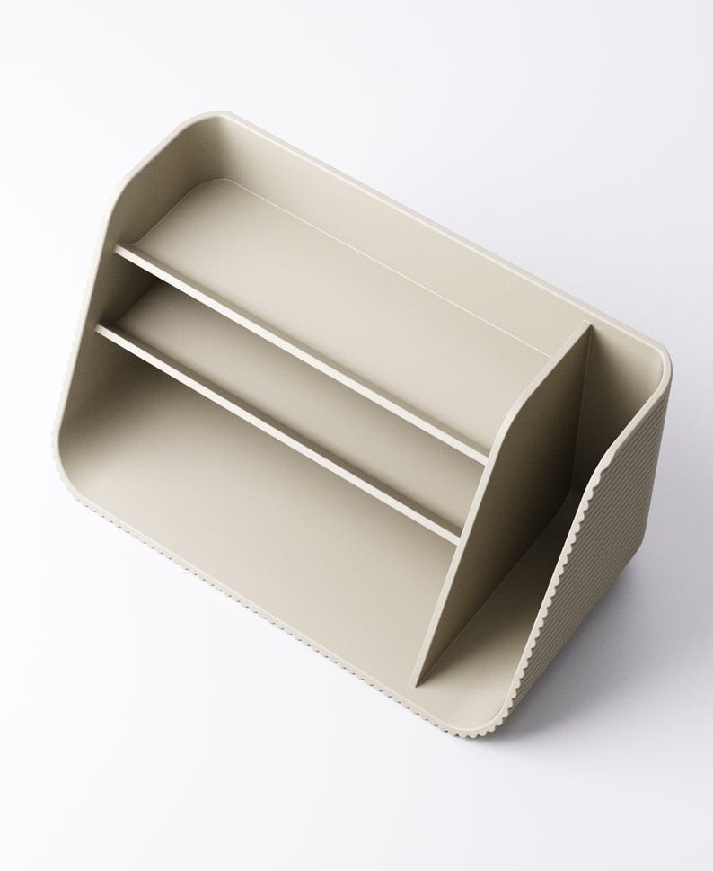 May include: A beige desk organizer with two shelves and a divided compartment. The organizer has a rounded shape with a textured side and a smooth, matte finish. It is designed to hold office supplies.