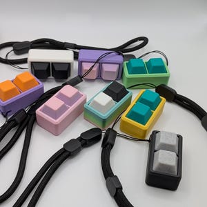 May include: A collection of colorful keycap pendants with black lanyards. The keycaps are in various colors, including white, black, purple, orange, pink, teal, and yellow. Each pendant features two keycaps.