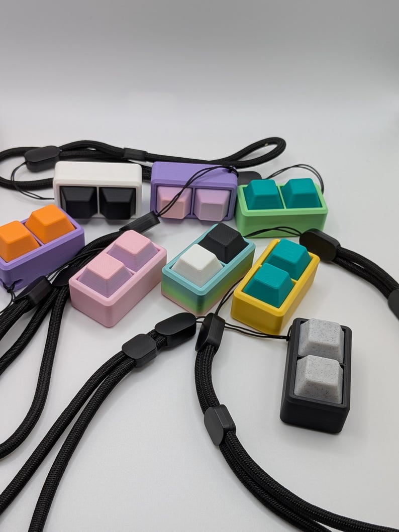May include: A collection of colorful keycap pendants with black lanyards. The keycaps are in various colors, including white, black, purple, orange, pink, teal, and yellow. Each pendant features two keycaps.