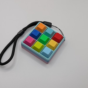 May include: A colorful, square fidget toy with a grid of pushable buttons in various colors, including orange, blue, green, and red. It has a black wrist strap attached. The toy is designed for sensory play and stress relief.