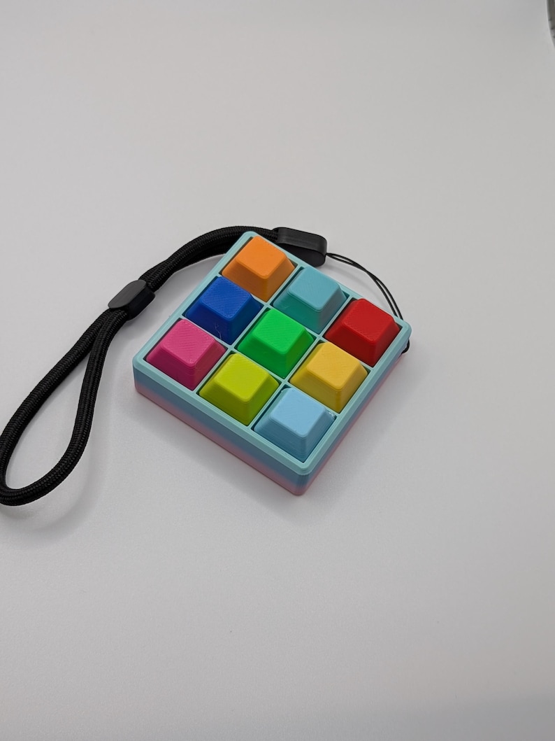 May include: A colorful, square fidget toy with a grid of pushable buttons in various colors, including orange, blue, green, and red. It has a black wrist strap attached. The toy is designed for sensory play and stress relief.