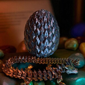 Op de afbeelding: Een gedetailleerd, metalen drakenei en een bijpassend drakenbeeldje. Het ei is donker met een textuur, schubachtig patroon. De draak is goud en bruin, met een gesegmenteerd lichaam en ingewikkelde details. De items rusten op een bed van gladde, kleurrijke stenen.