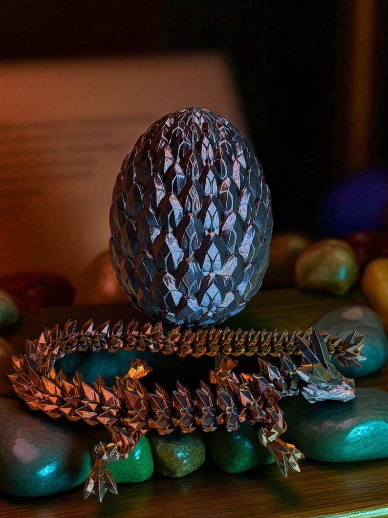 Op de afbeelding: Een gedetailleerd, metalen drakenei en een bijpassend drakenbeeldje. Het ei is donker met een textuur, schubachtig patroon. De draak is goud en bruin, met een gesegmenteerd lichaam en ingewikkelde details. De items rusten op een bed van gladde, kleurrijke stenen.