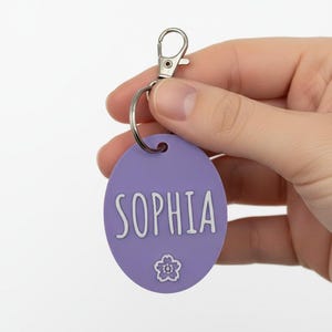 May include: A lavender oval keychain with the name "SOPHIA" in white script. A small flower graphic is below the name. The keychain has a silver clasp and ring.