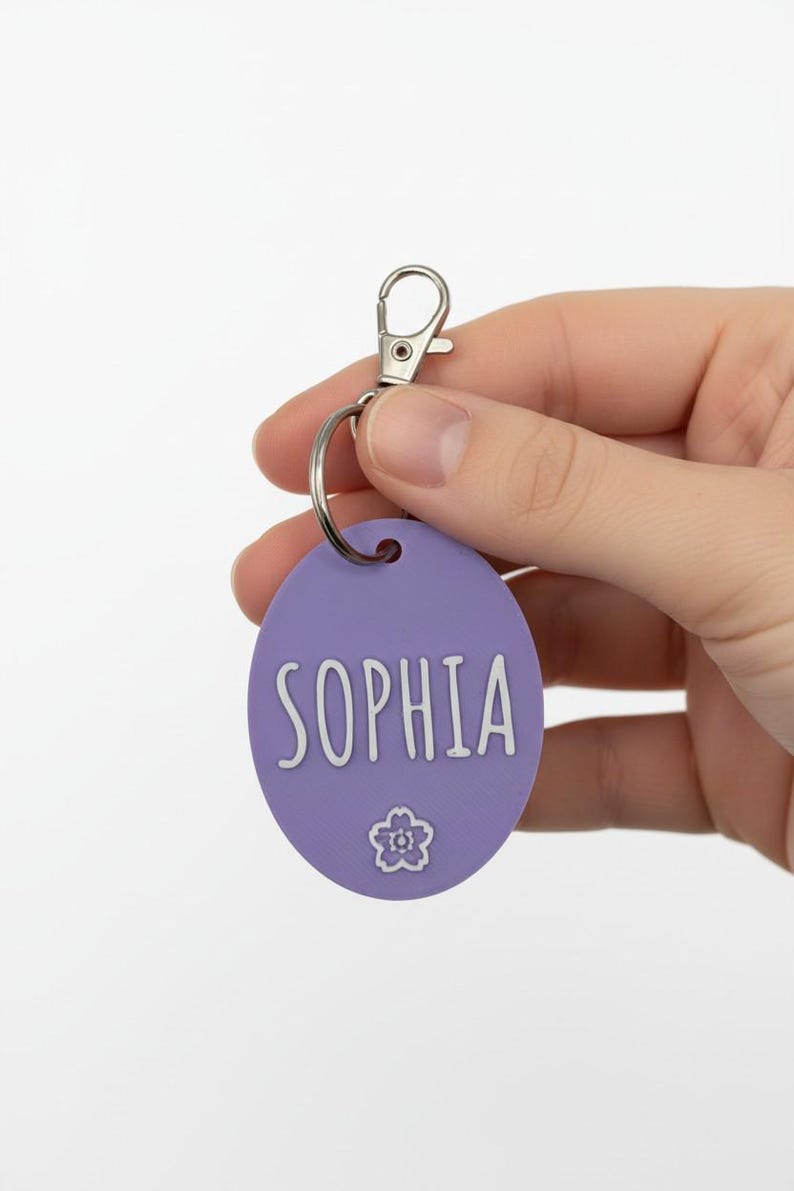 May include: A lavender oval keychain with the name "SOPHIA" in white script. A small flower graphic is below the name. The keychain has a silver clasp and ring.
