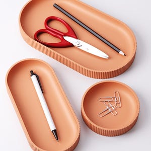 May include: A set of peach-colored desk organizers. The set includes an oval tray with red scissors and a pencil, another oval tray with a white pen, and a round dish with paper clips. The trays have a ribbed edge.