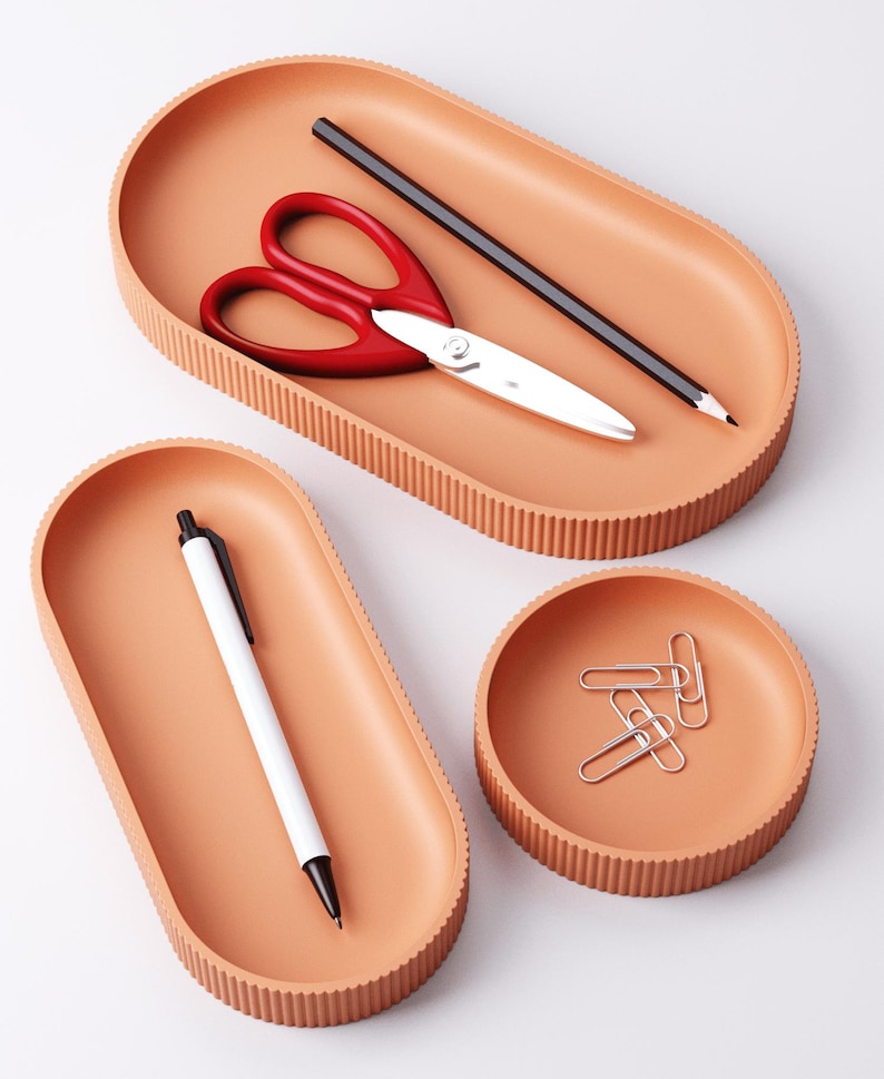 May include: A set of peach-colored desk organizers. The set includes an oval tray with red scissors and a pencil, another oval tray with a white pen, and a round dish with paper clips. The trays have a ribbed edge.