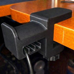 Puede incluir: Soporte negro para auriculares impreso en 3D, fijado a un escritorio de madera. La abrazadera tiene una parte superior curvada y una ranura para un cable de auriculares. El soporte est&aacute; dise&ntilde;ado para sujetar y organizar los auriculares.