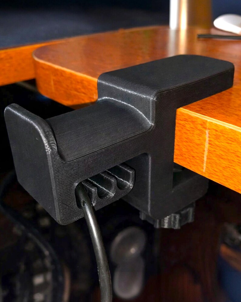 Puede incluir: Soporte negro para auriculares impreso en 3D, fijado a un escritorio de madera. La abrazadera tiene una parte superior curvada y una ranura para un cable de auriculares. El soporte est&aacute; dise&ntilde;ado para sujetar y organizar los auriculares.