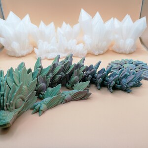 May include: A collection of 3D-printed sculptures. The top row features white, crystalline formations. Below, a teal and purple dragon-like sculpture is displayed alongside a circular, plant-like design. The objects are set against a neutral background.