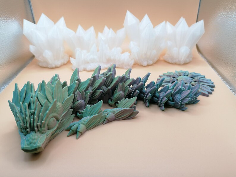 May include: A collection of 3D-printed sculptures. The top row features white, crystalline formations. Below, a teal and purple dragon-like sculpture is displayed alongside a circular, plant-like design. The objects are set against a neutral background.