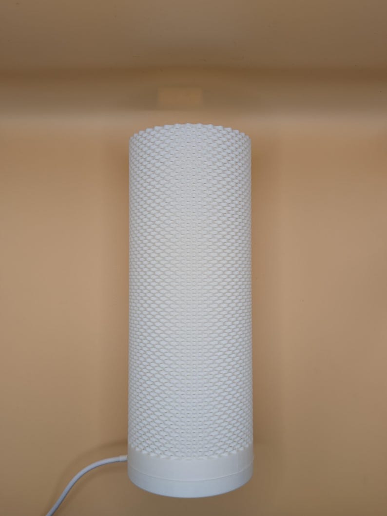K&ouml;nnte beinhalten: Eine wei&szlig;e zylindrische Lampe mit einer strukturierten, gepunkteten Oberfl&auml;che. Die Lampe hat einen soliden Sockel und ein Netzkabel. Der Hintergrund ist einfarbig beige. Die Lampe ist ein modernes Design.