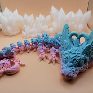 May include: A fantasy-themed sculpture of a dragon with a blue and pink gradient color scheme. The dragon has detailed features, including feathers, hearts, and clawed feet. Several white crystal formations are in the background.