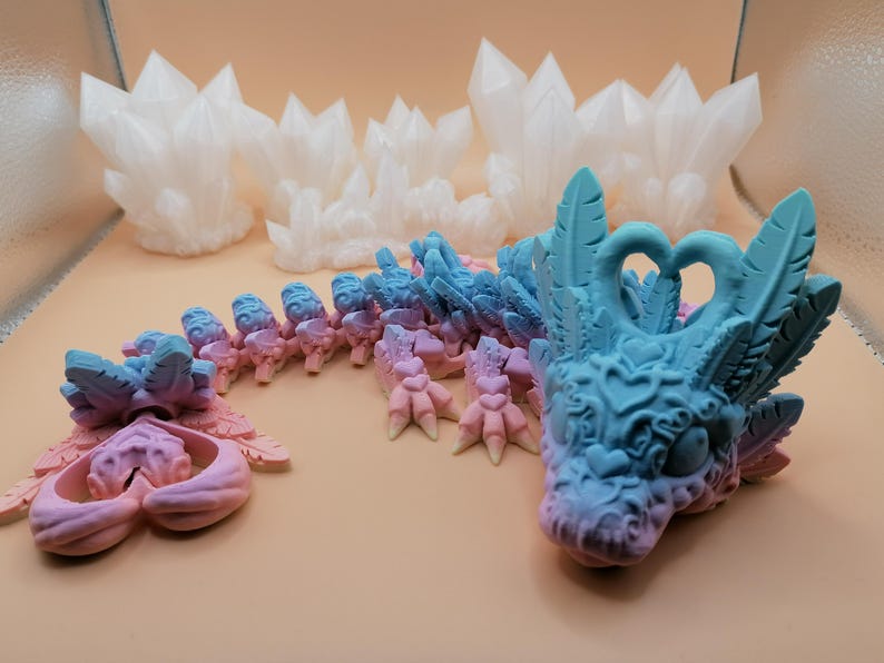 May include: A fantasy-themed sculpture of a dragon with a blue and pink gradient color scheme. The dragon has detailed features, including feathers, hearts, and clawed feet. Several white crystal formations are in the background.