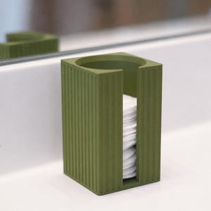 May include: A rectangular olive green cotton pad dispenser featuring a vertical ribbed texture on the exterior. The container has an open top and a vertical slot on the front, revealing a stack of white circular cotton pads stored inside.