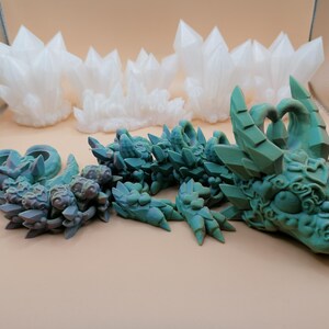 Op de afbeelding: Een gedetailleerde afbeelding toont een turquoise drakenbeeldje met ingewikkelde details, naast verschillende witte kristalformaties. De draak is in een ontspannen positie, met zijn hoofd en lichaam zichtbaar. De kristallen zijn gegroepeerd, wat een contrasterend element toevoegt.
