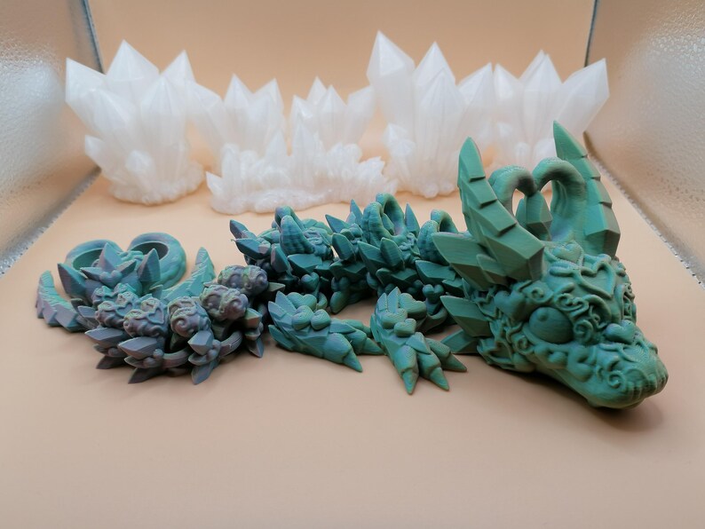 Op de afbeelding: Een gedetailleerde afbeelding toont een turquoise drakenbeeldje met ingewikkelde details, naast verschillende witte kristalformaties. De draak is in een ontspannen positie, met zijn hoofd en lichaam zichtbaar. De kristallen zijn gegroepeerd, wat een contrasterend element toevoegt.