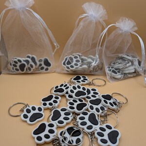 May include: White and black paw print keychains with silver keyrings. Some keychains are in white organza bags with drawstring closures. The keychains are scattered on a light brown surface.