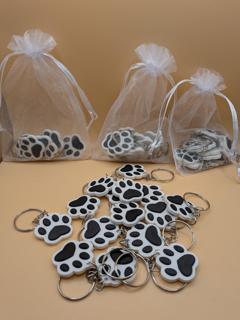 May include: White and black paw print keychains with silver keyrings. Some keychains are in white organza bags with drawstring closures. The keychains are scattered on a light brown surface.