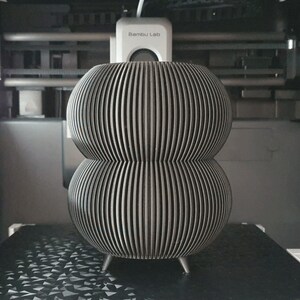 May include: A gray, 3D-printed decorative object with a stacked, bulbous design. The object features vertical, parallel lines and rests on three small legs. The item is inside a 3D printer.