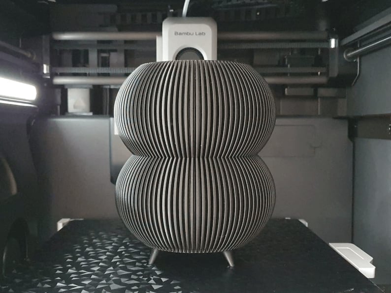 May include: A gray, 3D-printed decorative object with a stacked, bulbous design. The object features vertical, parallel lines and rests on three small legs. The item is inside a 3D printer.
