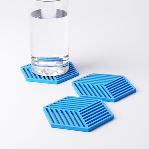 Pu&ograve; includere: Un bicchiere d'acqua &egrave; appoggiato su un sottobicchiere esagonale blu. Sono visibili anche altri due sottobicchieri blu. I sottobicchieri presentano un design geometrico con linee parallele. Lo sfondo &egrave; bianco.