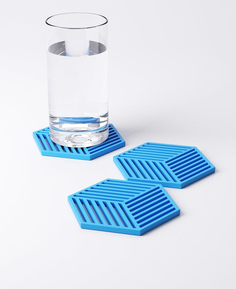 Pu&ograve; includere: Un bicchiere d'acqua &egrave; appoggiato su un sottobicchiere esagonale blu. Sono visibili anche altri due sottobicchieri blu. I sottobicchieri presentano un design geometrico con linee parallele. Lo sfondo &egrave; bianco.
