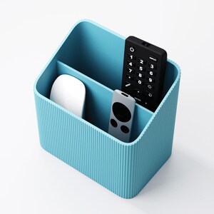 Puede incluir: Un organizador de escritorio azul claro con dos compartimentos. Un compartimento contiene un rat&oacute;n de ordenador blanco, mientras que el otro contiene un mando a distancia negro y un mando a distancia plateado. Dise&ntilde;ado para guardar material de oficina.